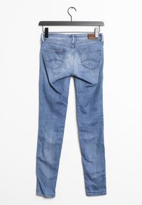 Jeans de denim azul colgados de un perchero negro. Presentan un corte ajustado, costuras contrastantes, dos bolsillos traseros y una etiqueta de cuero en la cinturilla.