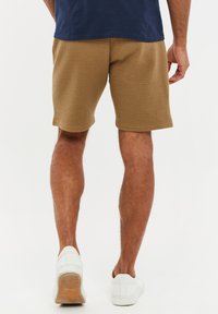 Homme portant un short brun texturé, une chemise marine et des baskets blanches, debout sur un fond blanc, vu de dos.