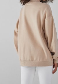 Beige oversize sweatshirt med hög hals, ribbad fåll och sänkta axlar, gjord av mjukt tyg. Kombinerad med vita byxor.