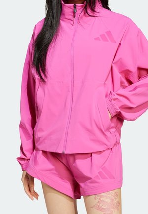 Woman wearing bright pink Adidas zip-up jacket and matching shorts, with a visible dragon tattoo on left thigh.