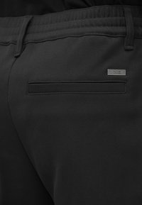 Pantaloni negri dintr-un material neted, cu un buzunar posterior cu deschidere orizontală și o etichetă din metal a mărcii. Talie elastică în partea de sus.
