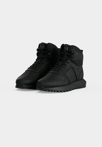 Ζευγάρι μαύρα sneakers ψηλού τύπου με υφή στις σόλες, κορδόνια μπροστά και επενδυμένο πέλμα αστραγάλου σε απλό φωτεινό φόντο.