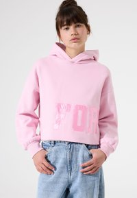 Sudadera corta rosa hecha de tejido suave, con un estampado brillante de "FOR" en la parte delantera, con capucha y puños elásticos. Lleva unos jeans de color azul claro.