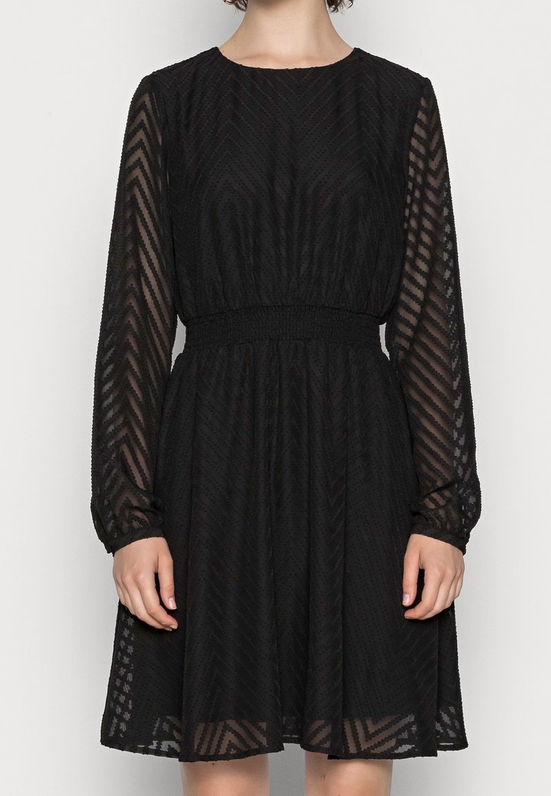 Svart klänning med långa, genomskinliga ärmar och en åtsittande smockad midja. Texturerad tyg har ett subtilt sicksackmönster. Utsvängd kjol.