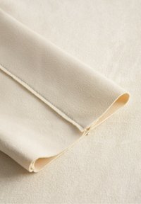 Tissu beige clair avec une texture douce et lisse, plié au bord, montrant un ourlet proprement cousu et un éclat subtil.