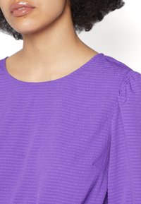 Top roxo com um tecido texturizado e finas riscas horizontais, decote arredondado e mangas curtas bufantes. Acabamento liso e leve.