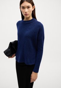 Navyblauer, flauschiger Strickpullover mit hohem Kragen, langen Ärmeln, lockerer Passform und leicht verkürztem Saum, kombiniert mit einer schwarzen Clutch.
