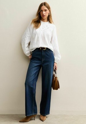 Blouse blanche texturée avec manches bouffantes et empiècement détaillé. Jean bleu foncé à jambes larges, bottines marron clair et sac à main marron.