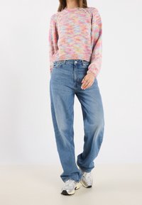 Femme portant un pull en maille pastel multicolore, un jean bleu ample et des baskets blanches, debout devant un fond blanc uni.