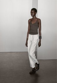 Bruine tanktop, witte linnen broek, zwarte sandalen. Accessoires omvatten een armband. Eenvoudig, aansluitend ontwerp met neutrale kleuren.
