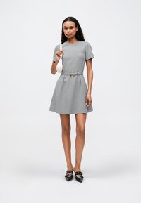 Γκρίζο καρό φόρεμα με κοντά μανίκια, εφαπτόμενη μέση και ζώνη. Φοριέται με μαύρες μυτερές τουαλέτες και κρατάει μια μικρή λευκή τσάντα.