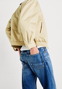 Person som bär en beige jacka med resår i midjan och blå denimjeans, med händerna i bakfickorna, mot en enfärgad vit bakgrund.