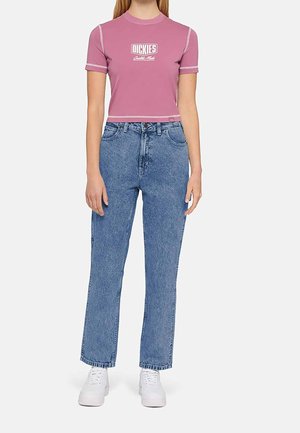 Woman wearing a pink crop top with "Dickies" logo, high-waisted blue jeans, and white sneakers, standing against a plain background.