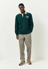 Dunkelgrünes langärmliges Polohemd mit beigem Kragen, versehen mit einem Logo. Getragen mit hellbeigen Cordhosen und braunen Sneakers.