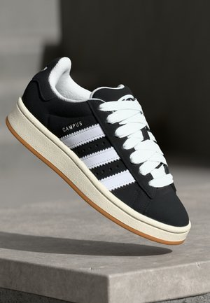 Baskets Adidas Campus noires avec des bandes blanches et des lacets, semelle beige, inclinées comme en plein vol au-dessus d'une surface en pierre.