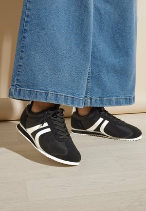 Black sneakers with a white side design, made from a smooth material, featuring round laces and a textured white sole.
