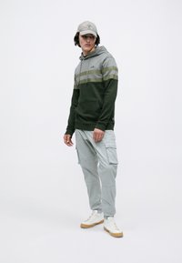Młody mężczyzna ubrany w zielono-szary hoodie, jasnoszare spodnie cargo, białe sneakersy i jasny czapkę baseballową, stojący na białym tle.
