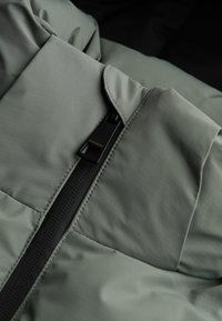Grijze pufferjack met een gladde textuur, voorzien van een zwarte rits en logo-tag. Gestikte, gequiltte delen verbeteren de isolatie.