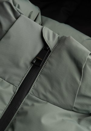 Szara kurtka puchowa o gładkiej fakturze, z czarnym suwakiem i metką z logo. Szyć w quilted sekcjach zwiększa izolację.