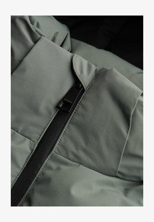 Graue Pufferjackenstoff mit glatter Textur, ausgestattet mit einem schwarzen Reißverschluss und einem Logotag. Genähte gesteppten Abschnitte verbessern die Isolierung.