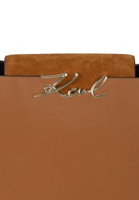 Brązowa skórzana torba o gładkim wykończeniu, z beżowym zamszowym wierzchem i metalicznym złotym akcentem z logo "Karl" z przodu.