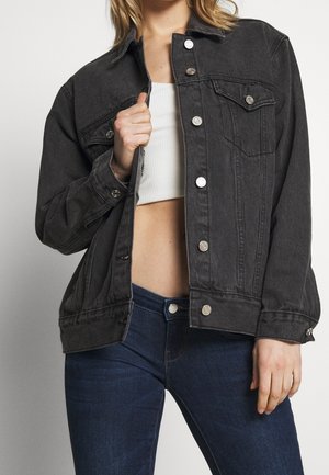 Femme portant une veste en jean boutonnée gris foncé sur un crop top blanc et un jean bleu foncé, tenant la veste ouverte d'une main.