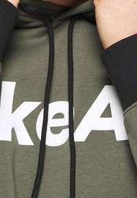 Sudadera de algodón verde con un logo blanco, puños negros y cordones. La tela tiene una textura suave y un diseño de ajuste relajado.