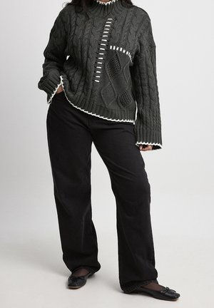 Kobieta w ciemnoszarym swetrze warkoczowym z białym wykończeniem i czarnych szerokich spodniach, stojąca z jedną ręką w kieszeni.