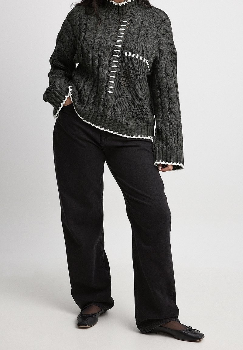 Femme portant un pull gris foncé à torsades avec bordure blanche et un pantalon large noir, debout avec une main dans la poche.