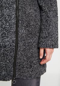 Cappotto grigio con texture e materiale peloso. Presenta una cerniera anteriore e un orlo arrotondato. Inquadratura ravvicinata che mostra la manica e la mano che afferra il tessuto.