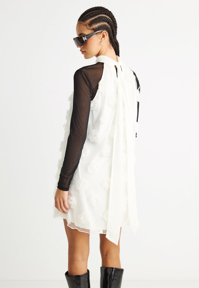 Kobieta z zaplecionymi włosami, nosząca czarne okulary przeciwsłoneczne, przezroczystą czarną bluzkę z długim rękawem pod białą sukienką o kwiatowej fakturze i długimi paskami z tyłu, czarne buty.