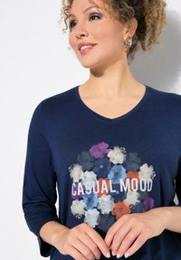 Maglietta a V blu navy realizzata in tessuto morbido, caratterizzata da una stampa floreale in vari colori con il testo "CASUAL MOOD" in bianco.