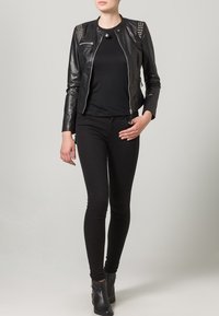 Černá kožená bunda s cvočkovanými detaily na ramenou, kombinovaná s černým topem a úzkými černými kalhotami. Modelka má na sobě černé kotníkové boty.