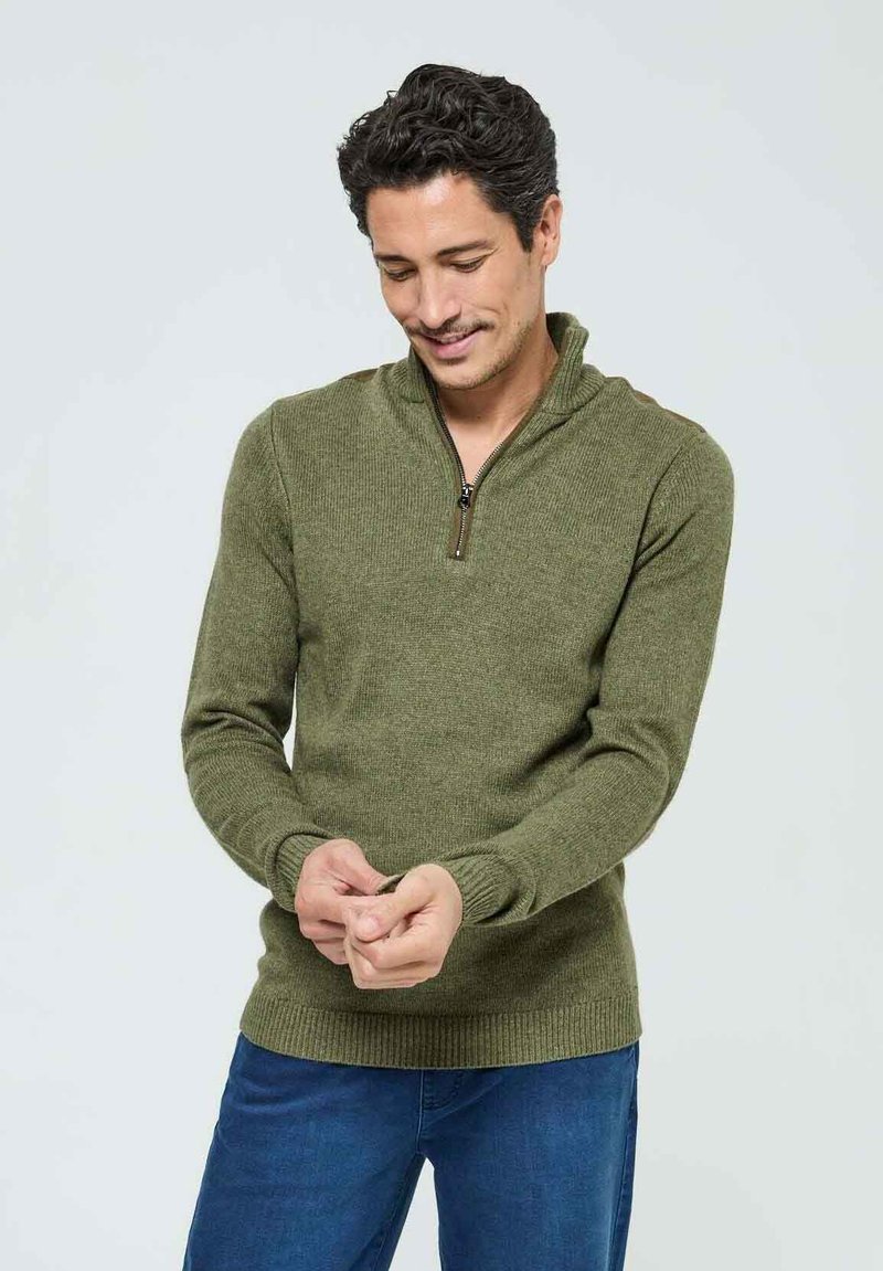 Pull en maille vert avec un col zippé mi-hauteur, des poignets et un ourlet côtelés. Porté sur un jean bleu, avec un tissu texturé.