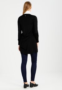Schwarze Langarm-Tunika aus strukturierter Stoff mit seitlichen Schlitzen, kombiniert mit dunkelblauen Slim-Fit Jeans und schwarzen flachen Schuhen. Weißer Kragen ist sichtbar.