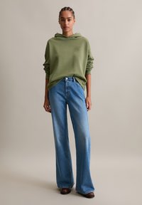 Felpa con cappuccio verde oliva con logo frontale, abbinata a jeans a gamba larga blu chiaro. Tessuto morbido e vestibilità rilassata dallo stile casual.