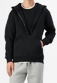 Sudadera negra con cremallera, bolsillo frontal, capucha ajustable y puños de canalé. Diseño casual, combinada con pantalones de chándal grises. Textura de tela suave.