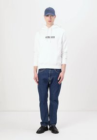 Hvid hættetrøje med logo, blå baseballcap, mørkeblå løse jeans og sorte sko. Simpelt design med minimal detaljering.