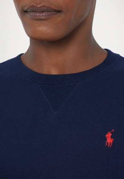 Marineblå strikkegenser med ribbestrikket hals og mansjetter. Har en liten rød polo spiller-logo på venstre bryst. Glatt tekstur, uformelt design.