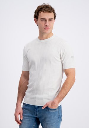 Jeune homme aux cheveux bouclés portant un pull côtelé blanc à manches courtes et un jean bleu, debout avec une main dans la poche.