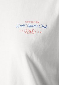 T-shirt bianco con testo rosso e blu. Il testo recita: "NEW HAVEN Gant Sports Club USA 1949." Texture liscia con un design casual.