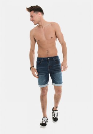 Shorts en denim bleu foncé avec un ourlet retroussé, dotés de cinq poches et d'une fermeture à bouton, assortis à des baskets noires.
