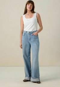 Lichtblauwe high-waisted jeans met een wijduitlopende pasvorm, omgeslagen zooms, gecombineerd met een aansluitend wit geribbeld tanktop en patroonaccenten op de schoenen.