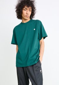 Teal cotton t-shirt with short sleeves and a crew neckline; features a small white logo on the chest. Worn with black athletic pants.
