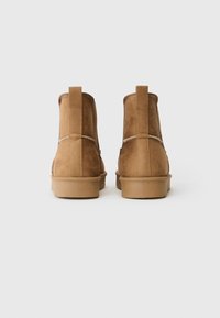 Pair of tan suede ankle boots with pull tabs, visible stitching, and flat rubber soles, shown from the back on a plain background.