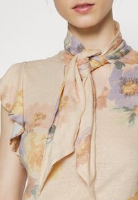 Närbild på en person som bär en beige blommig topp med en matchande blommig sjal knuten runt halsen.