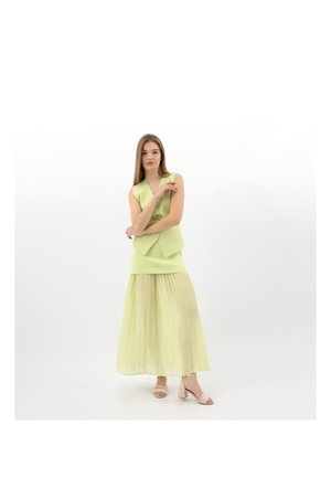 Donna in piedi con le braccia incrociate, indossa un gilet senza maniche verde chiaro e una gonna lunga plissettata coordinata, con tacchi beige a blocco.