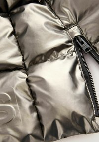 Detail einer metallischen Pufferjacke mit gestepptem Texture, glänzender Oberfläche und dunklem Reißverschluss mit einem Marken-Zugtab.