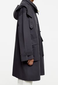 Manteau léger gris foncé avec capuche, grandes poches avant, fermetures à boutons et pressions, et poignets ajustables. Texture lisse et coupe décontractée.