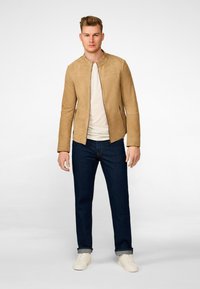 Tan suède jas met een kraag, ritszakken en lange mouwen, gecombineerd met donkerblauwe jeans en lichte sneakers.